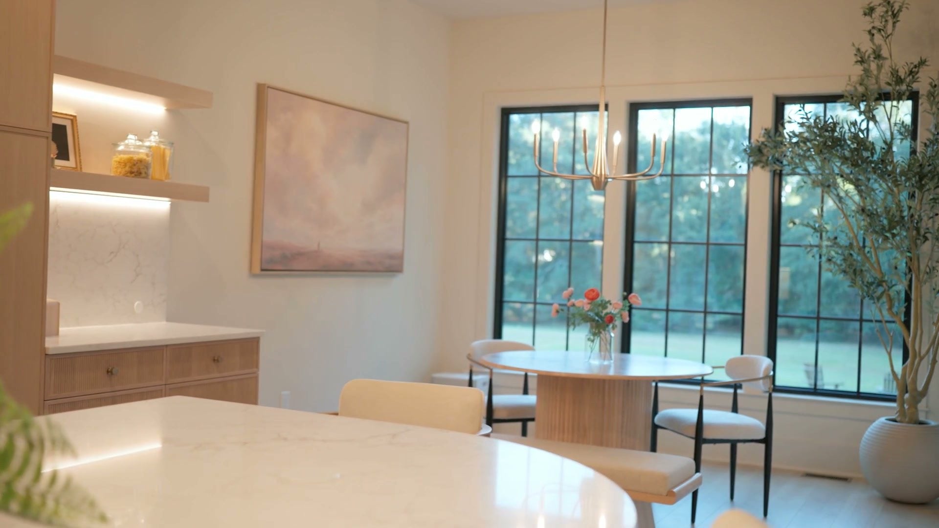 The dining area: a round oak table in front of tall black-framed windows, open shelving and the kitchen counter in the foreground