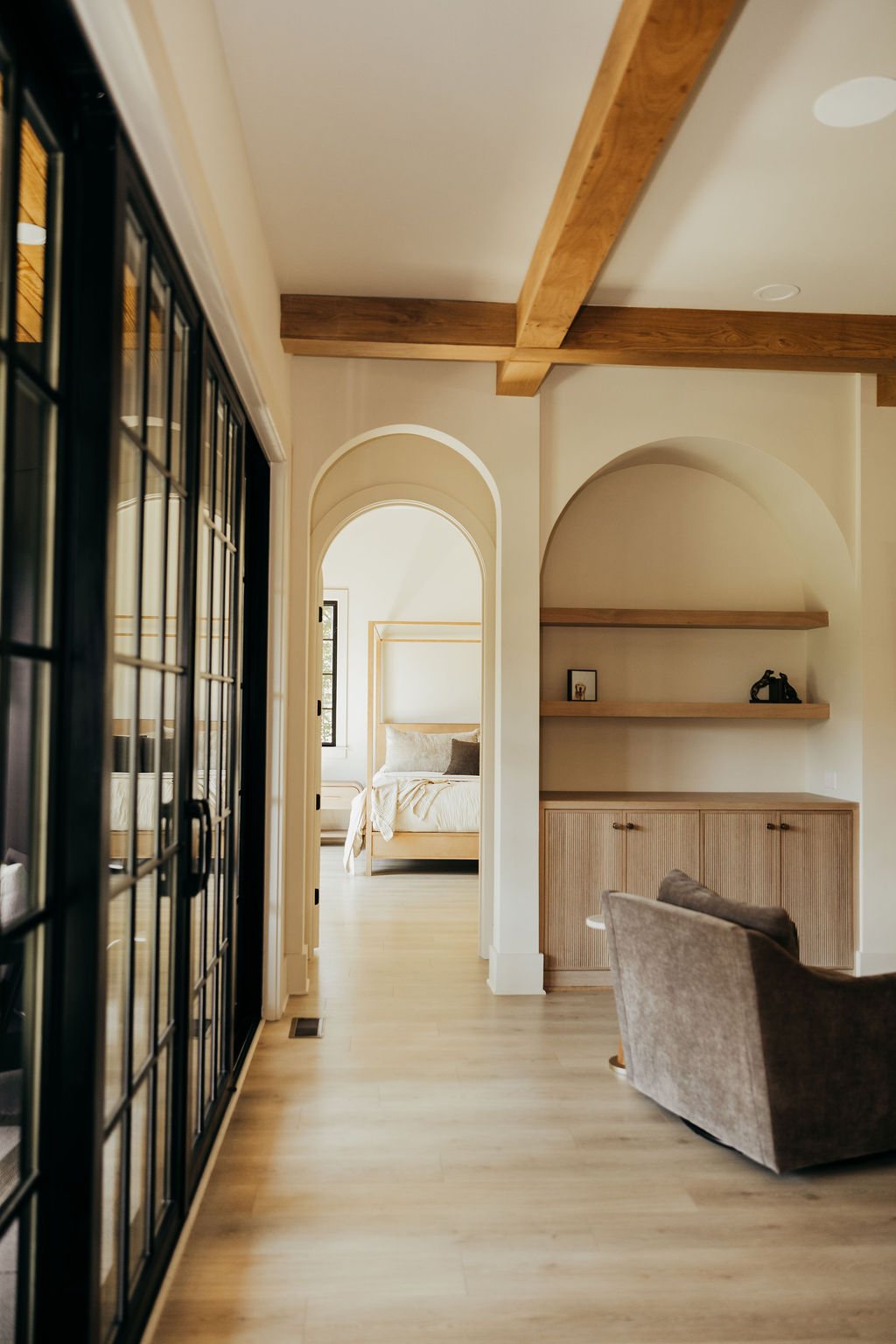 The hallway: a run of arched openings past open shelving, steel-framed glass on one side, oak beams above