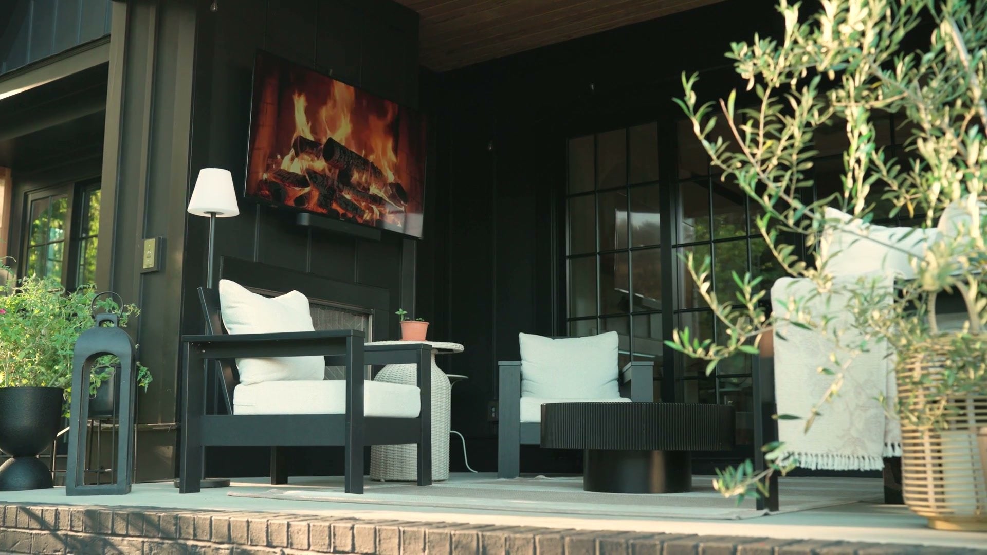 The covered back porch: armchairs and an olive tree under a wood ceiling, the fireplace lit on the dark wall